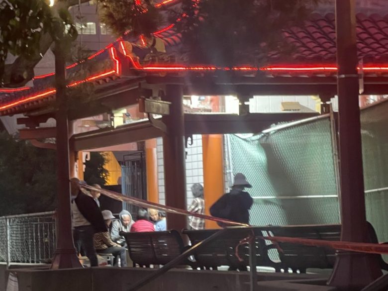 A group of people sit and stand near benches under a pavilion at night, with a red neon-lit roof and a green construction fence nearby.