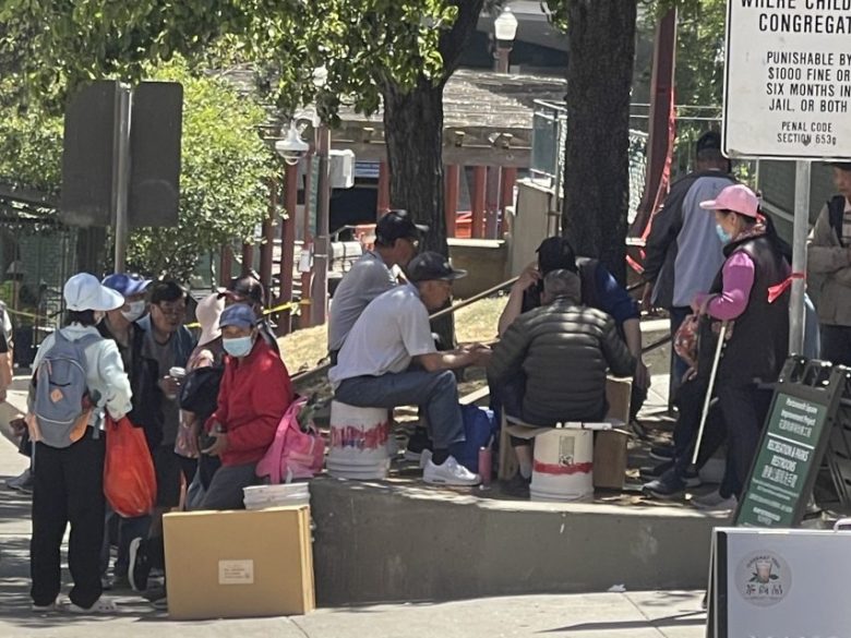 A group of people gather outdoors near a sidewalk, some sitting on buckets and others standing, with bags and boxes around them under trees and a street sign.