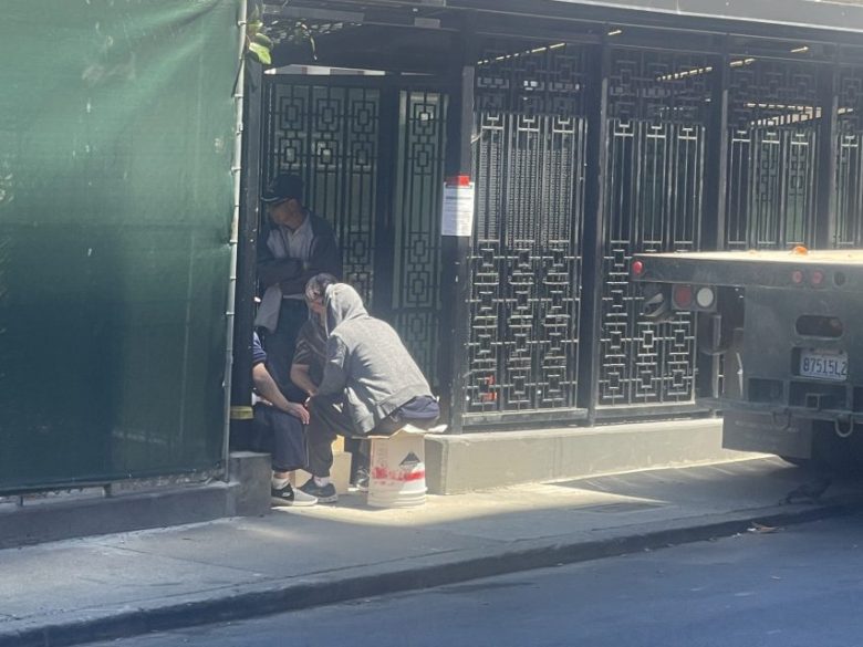Four people sit and crouch together on a sidewalk next to a building with decorative metalwork, partially shaded by green fencing, with a truck parked nearby.