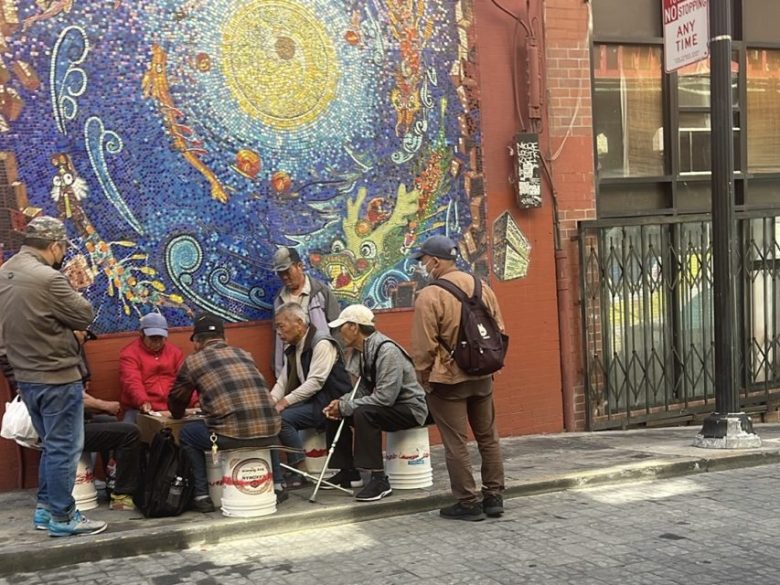 A group of men sit and stand on a sidewalk playing a game on overturned buckets near a colorful mosaic mural on a brick wall.