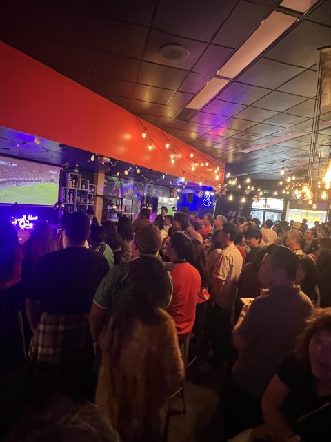 A crowd of people stands inside a dimly lit bar, watching a sports game on a large screen. String lights hang from the ceiling, creating a lively atmosphere.