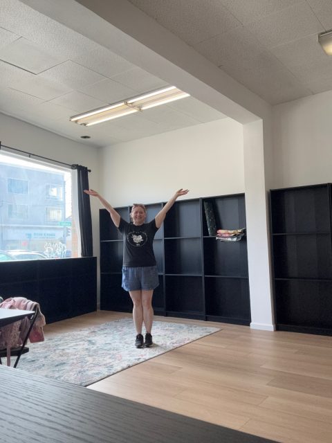 A person stands in the center of a mostly empty room with black shelves, a large window, and a rug, raising both arms and smiling at the camera.