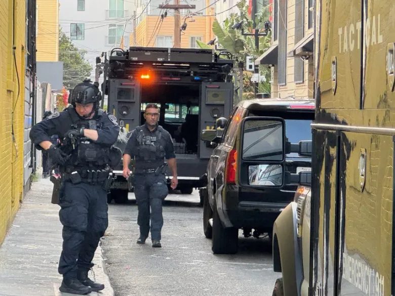 Three police officers in tactical gear stand near armored vehicles in an urban alleyway, with one officer adjusting equipment on his vest.
