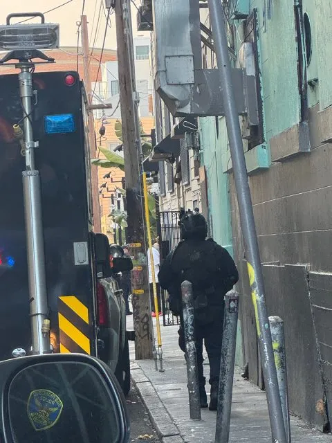A person in tactical gear walks down a narrow alley beside utility poles, with a black vehicle and equipment nearby.