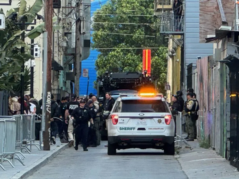 A group of law enforcement officers and a sheriff’s vehicle are gathered in a narrow urban alleyway near metal barricades and buildings.