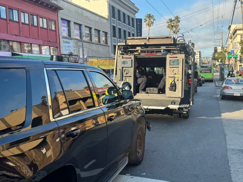 A police SWAT vehicle and a black SUV are parked on a city street, with officers visible inside the SWAT vehicle and pedestrians nearby.