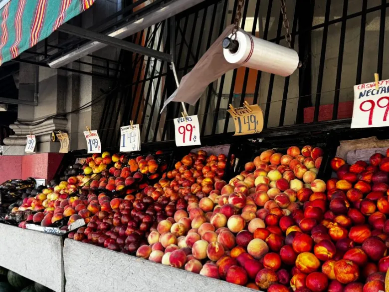 Display of peaches, nectarines, and plums at an outdoor market stand, each with handwritten price signs, under a striped awning and a hanging roll of produce bags.