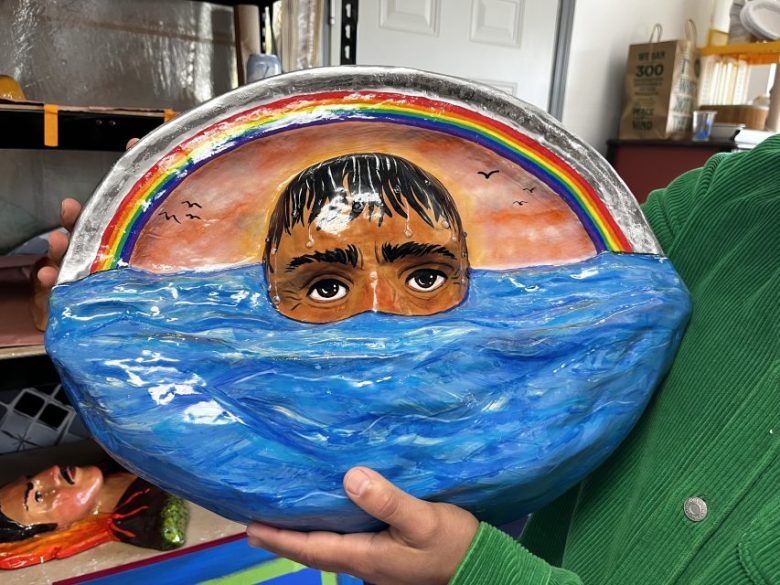 A person holds a ceramic artwork depicting a face partially submerged in blue water with a rainbow and birds in the background.