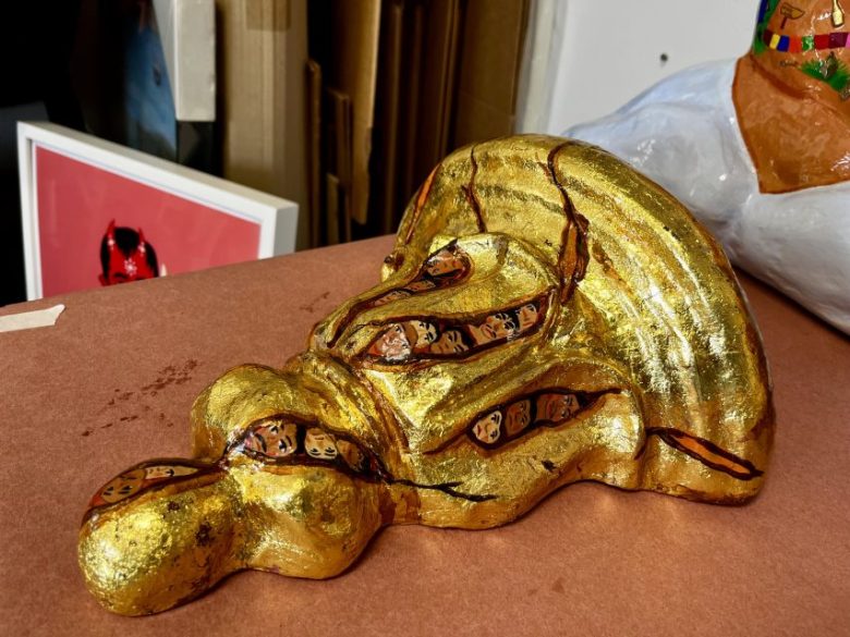 A gold-painted sculpted object with embedded faces rests on a brown surface, with framed artwork and other objects in the background.