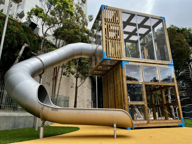 A modern playground structure with a tall wooden and glass climbing tower and a large metal tube slide on a yellow rubber surface.