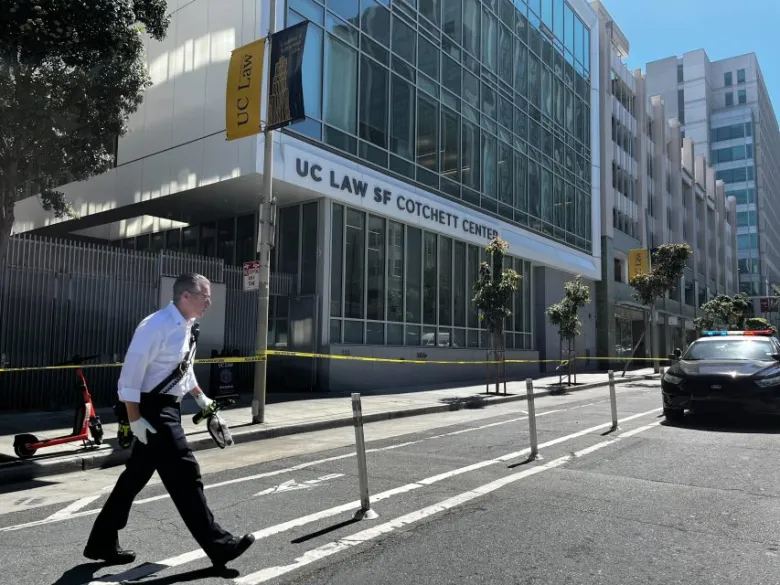 A man in business attire walks past yellow police tape outside the UC Law SF Cotchett Center in the Tenderloin after a recent shooting on a sunny day.