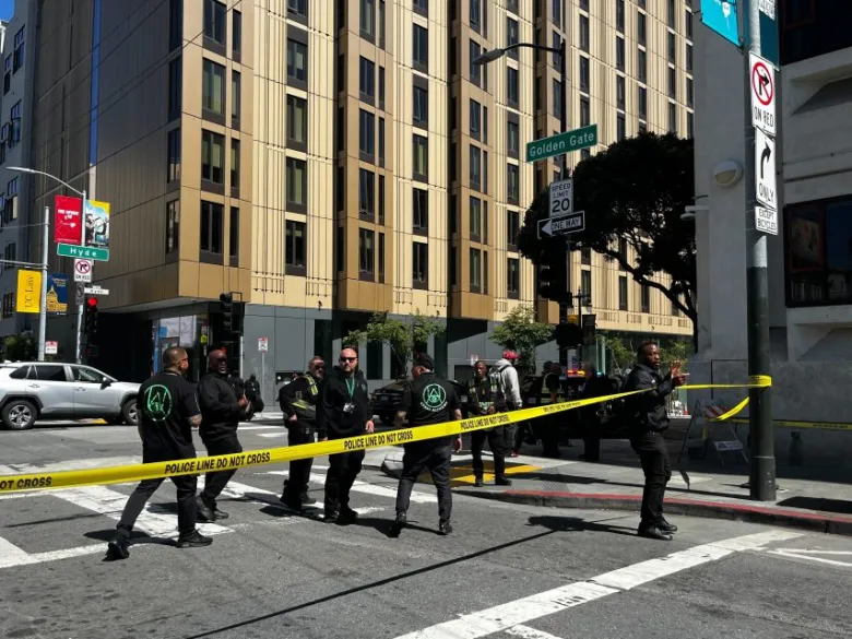 Police officers stand behind yellow caution tape at a city street intersection in the Tenderloin district, in front of a tall building on a sunny day, responding to a reported shooting.