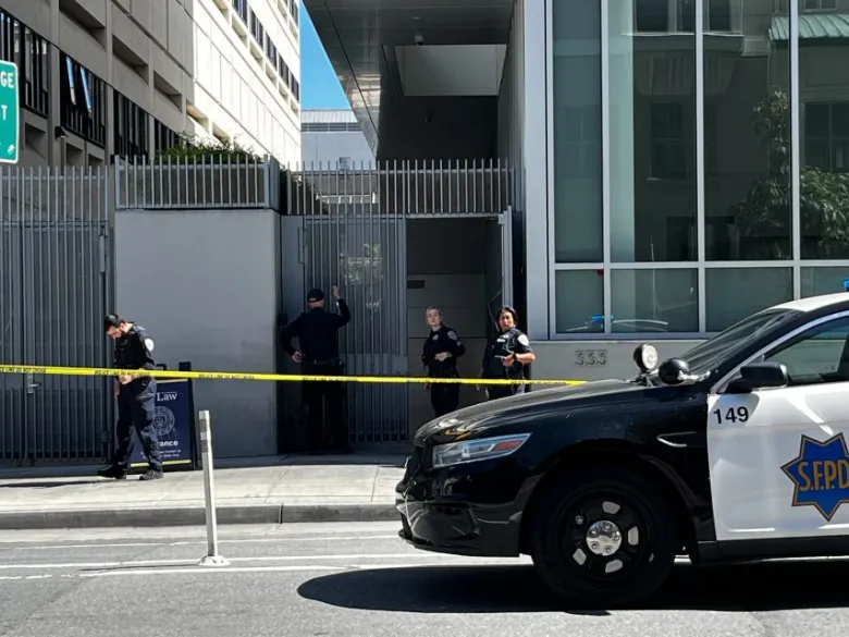 Police officers stand behind yellow caution tape outside a modern building in the Tenderloin, as a police car is parked in the foreground following a reported shooting.