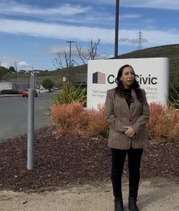 A woman stands in front of a company sign with rolling hills and a partly cloudy sky in the background.