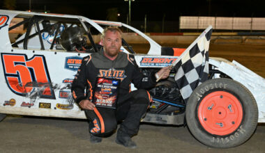 Racing driver kneels beside a white dirt late-model race car number 51, holding a checkered flag at night on the track.
