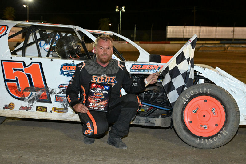 Racing driver kneels beside a white dirt late-model race car number 51, holding a checkered flag at night on the track.