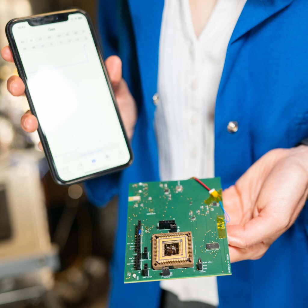 A person holds an iPhone in one hand and a green electronic board with the electronic nose attached in the other hand.