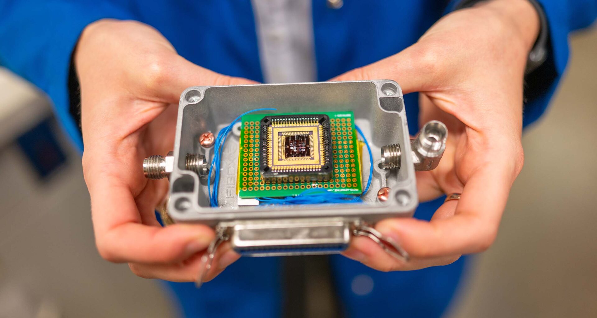 A pair of hands holds a small plastic box that contains a complex electronic chip.
