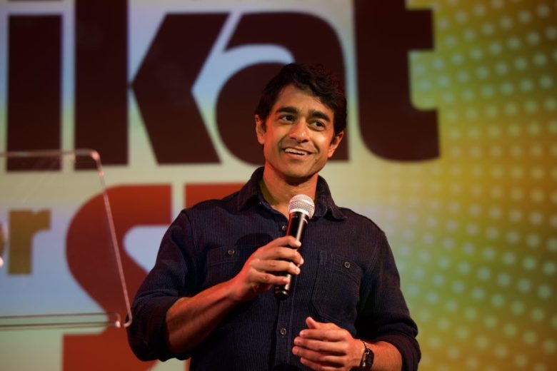 A man stands on stage holding a microphone, smiling, with a colorful background displaying large, partially visible text.