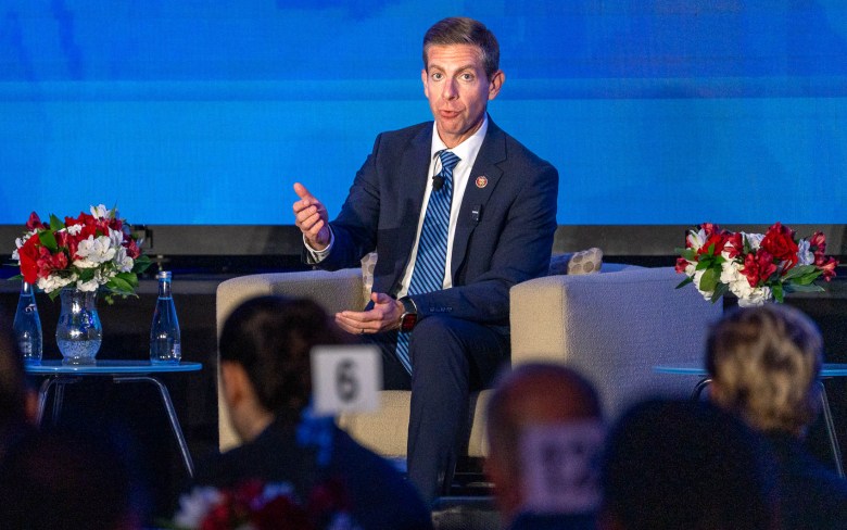 A man sits between two tables in an armchair speaking, while raising his right hand, with thumb up, to make a point.