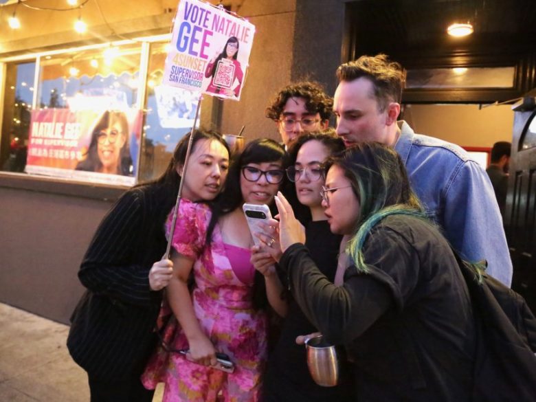 A group of six people stand close together outside, looking at a smartphone; one holds a “Vote Natalie Gee” campaign sign.