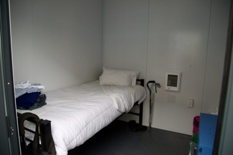 A small, plain room with a single bed, folded clothes on the bed, a cane, shoes on the floor, and a wall vent.