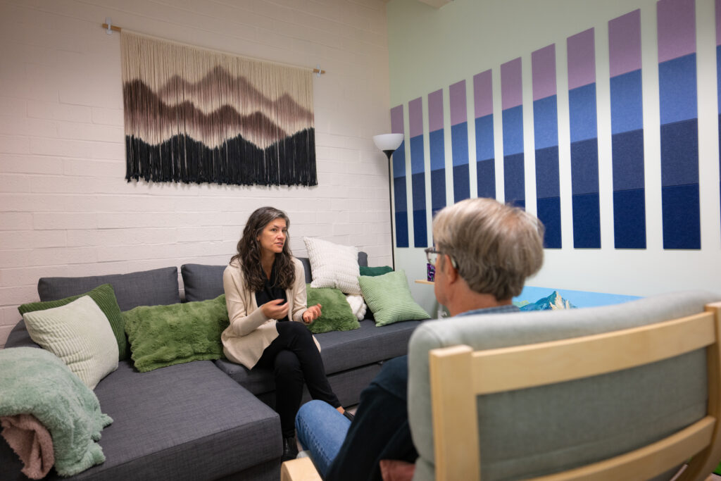 Two people sit across from one another in a brightly lit room that is furnished with a comfortable couch and chair and colorful art on the wall.