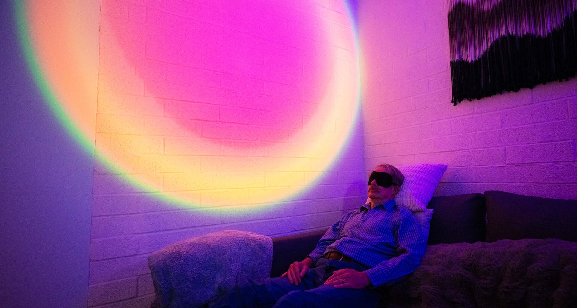 A man wearing an eye mask reclines on a couch in a darkened room. Behind him, the wall is illuminated with a circular, rainbow-colored light.