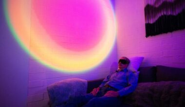 A man wearing an eye mask reclines on a couch in a darkened room. Behind him, the wall is illuminated with a circular, rainbow-colored light.