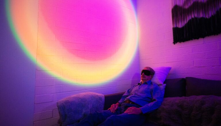 A man wearing an eye mask reclines on a couch in a darkened room. Behind him, the wall is illuminated with a circular, rainbow-colored light.
