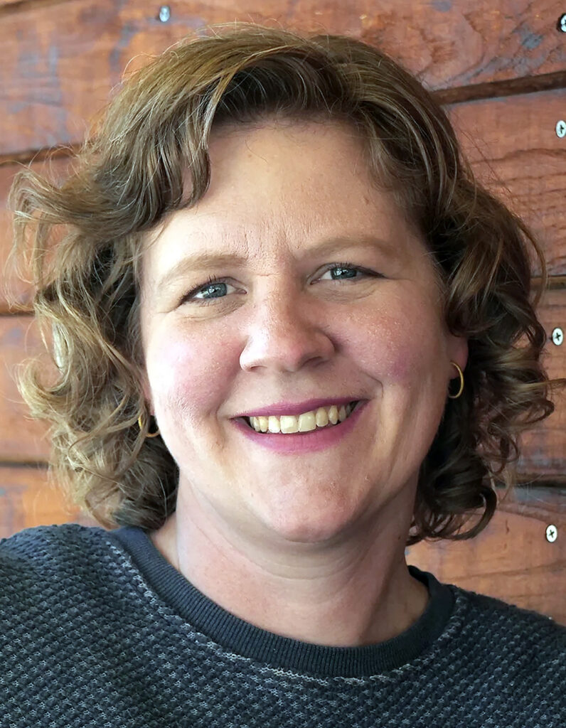 informal headshot of Miranda Dietz, smiling, against a backdrop of orange-red stained wood planks