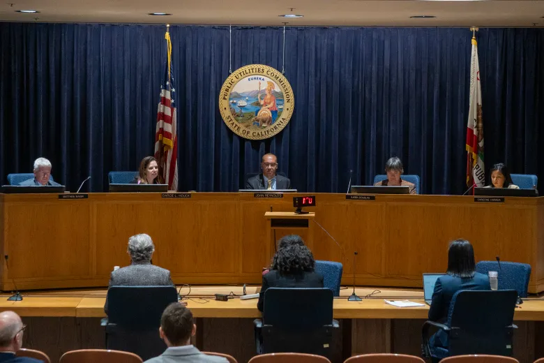 five people sit behind a large curved desk in a hearing room with American and California flags and the seal of the Public Utilities Commission behind them