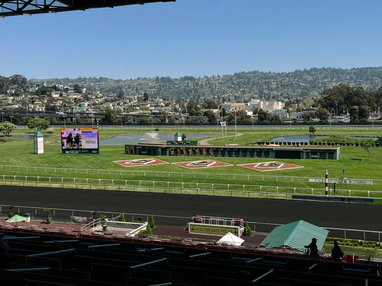 A view of a horse racing track from a grandstand seat, looking out onto a green infield and byond that to the East Bay Hills
