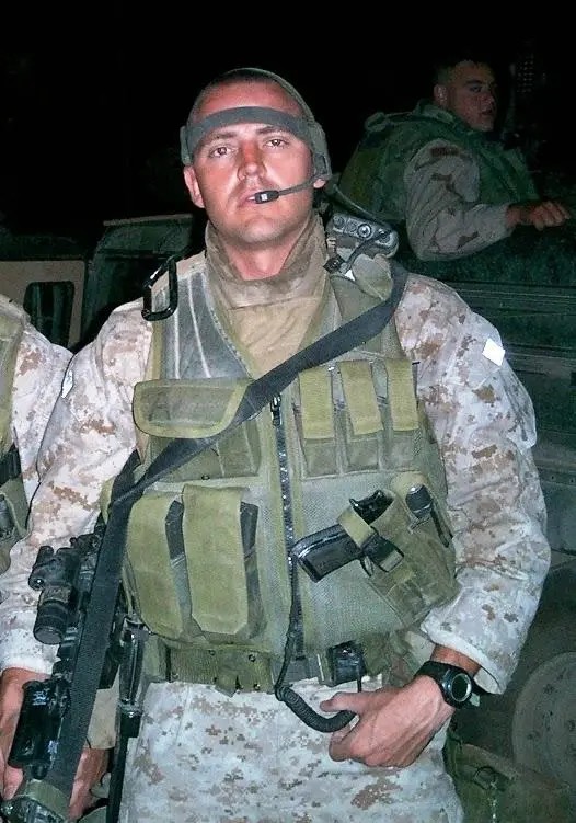 A soldier in camouflage gear stands at night, wearing a tactical vest and headset, exuding focus and readiness. Another soldier is visible in the background.