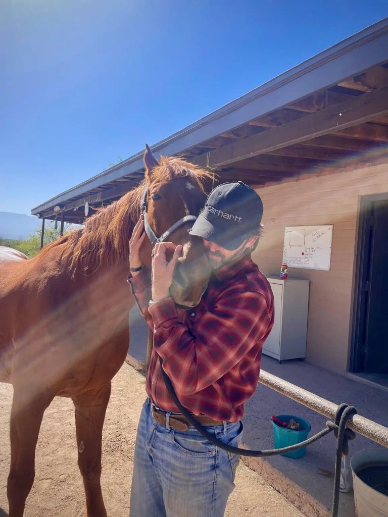 A person in a black cap and red plaid shirt kisses a chestnut horse in a stable, with a clear sky and wooden beams in the background.