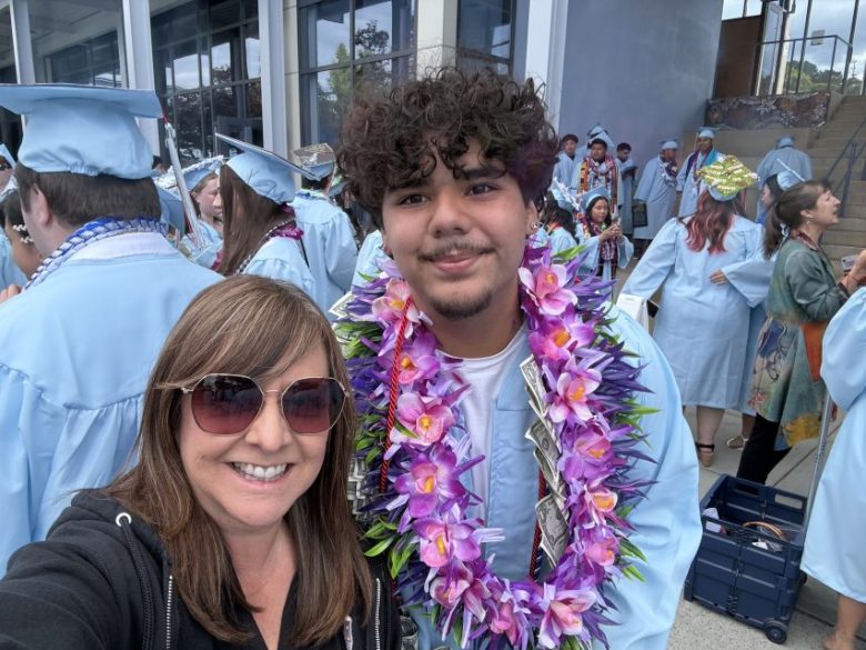 A woman in sunglasses takes a selfie with a graduate wearing a blue cap and gown and purple floral leis at an outdoor graduation ceremony. Other graduates are visible in the background.