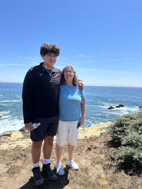 A young man and an older woman stand together on a rocky coastline with the ocean and blue sky in the background.