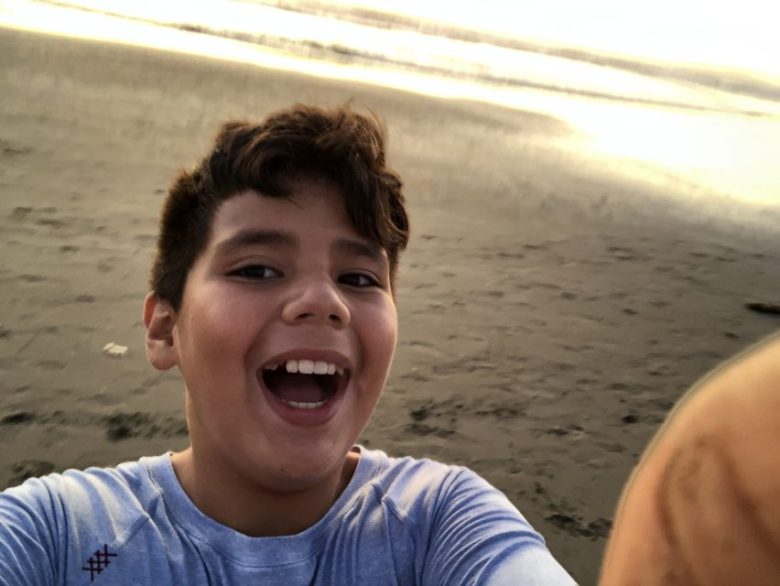 A young boy takes a selfie on a sandy beach at sunset, smiling with the ocean and waves visible in the background.