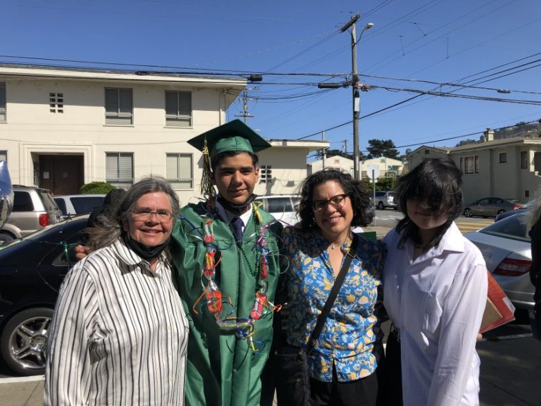 Four people stand together outdoors; one is wearing a green graduation cap and gown with leis, while the others are dressed in casual clothing. Buildings, cars, and power lines are visible behind them.