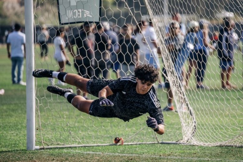 A soccer goalkeeper in mid-air attempts to block a shot, with spectators and players visible in the background.