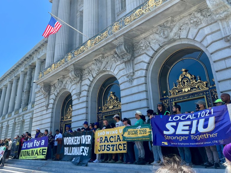 Protesters from SEIU and other orgs hold up signs on City Hall steps