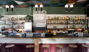 A modern bar with six stools, a long counter, shelves of bottles, a drink menu above taps, geometric ceiling art, and hanging spherical light fixtures.
