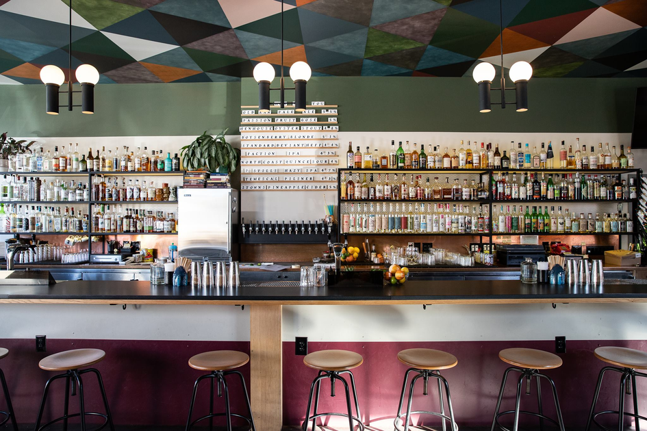 A modern bar with six stools, a long counter, shelves of bottles, a drink menu above taps, geometric ceiling art, and hanging spherical light fixtures.