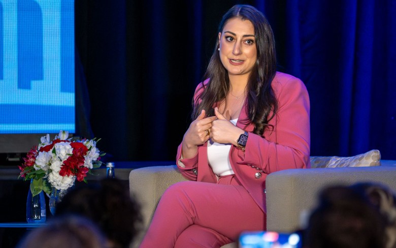 A woman sits beside a table with flowers. She is folding her hands in toward her chest while speaking.