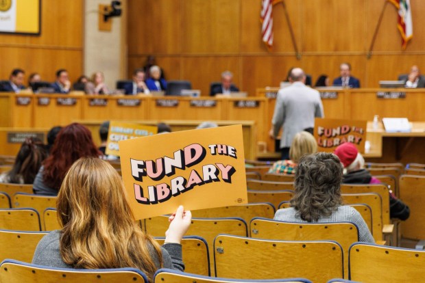Members of the public hold up signs in protest against library budget cuts during a budget review committee meeting at City Hall on Friday, June 5, 2026. (Kristian Carreon / The San Diego Union-Tribune)