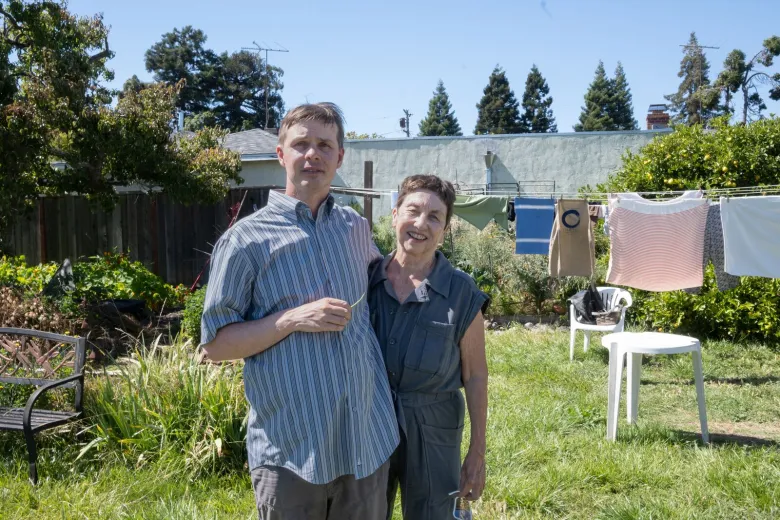 A man and woman stand arm-in-arm in a grassy lawn, with patio furniture, trees and laundry hanging on a clothesline behind them