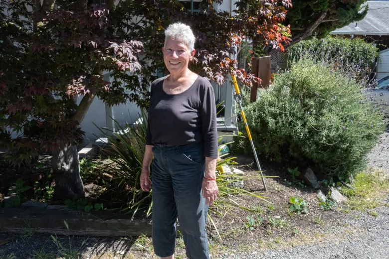 A woman stands on a gravel driveway, in front of a tree and a lavender bush