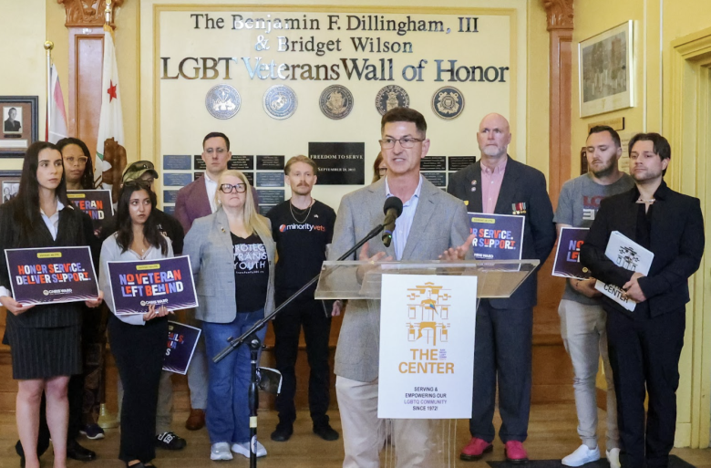 A group of people stand at a podium in front of an "LGBT Veterans Wall of Honor." The central speaker holds a microphone, while others hold signs with slogans like "Honor Service, Deliver Support" and "No Veteran Left Behind."