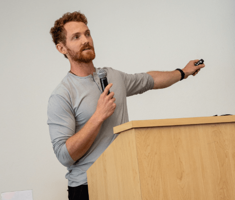 A man with curly hair speaks into a microphone while gesturing to a screen.