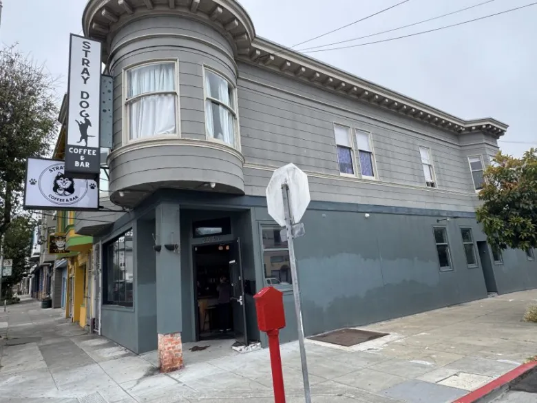 A gray two-story building on a street corner houses Stray Dog Coffee Bar, with signage visible above the entrance and a person standing inside the doorway.